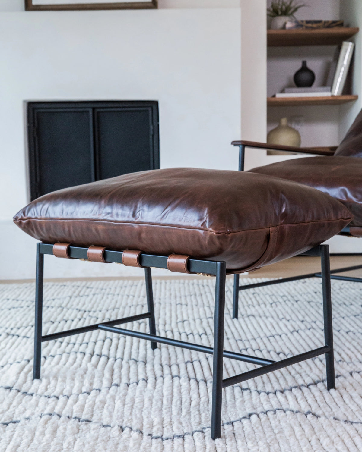 Close-up view of a Vail Ottoman in a cozy living room, with a plush brown leather cushion on a sleek black metal frame. The cushion’s craftsmanship is evident in the detailed stitching and piping, and it is supported by a series of cylindrical wood accents between the frame's legs. 