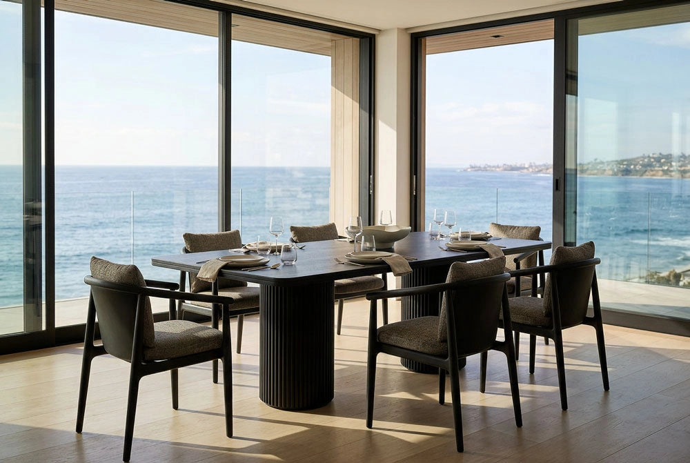 The Rio Dining Table with Dillon Dining Chairs by large windows with a view of the ocean.